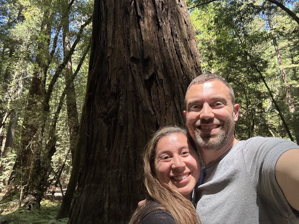 A couple smiling at the foot of an enormous redwood, sunlight coming through the canopy behind them
