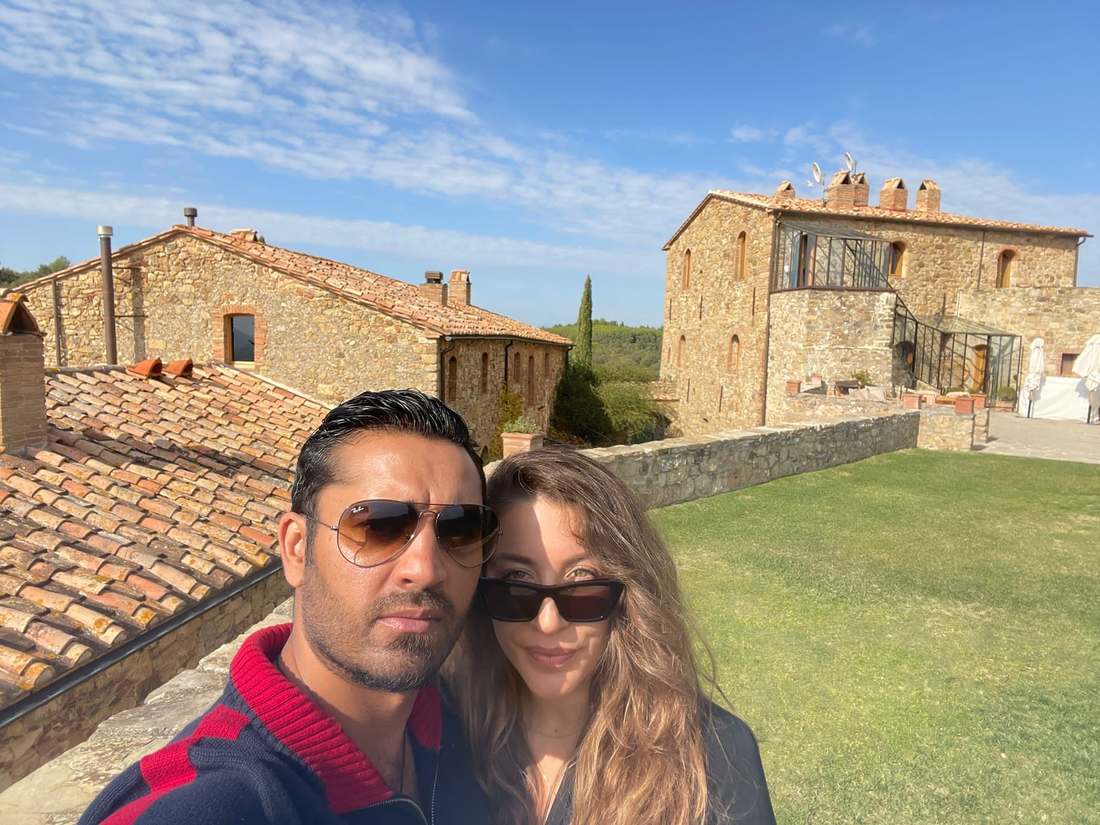 A couple in sunglasses in front of a stone farmhouse with terracotta roofs under a blue sky
