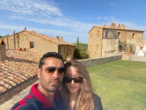A couple in sunglasses in front of a stone farmhouse with terracotta roofs under a blue sky